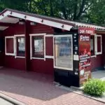 Kleines rotes Holzhaus mit weißen Fensterrahmen und Rollläden, davor ein Getränkeautomat mit rotem Aufsteller mit der Aufschrift "24H-THEKE" und Angeboten wie Grillartikel, Fleisch, Wurst, Konserven, Eier, umgeben von Pflanzen und einem gepflasterten Gehweg.
