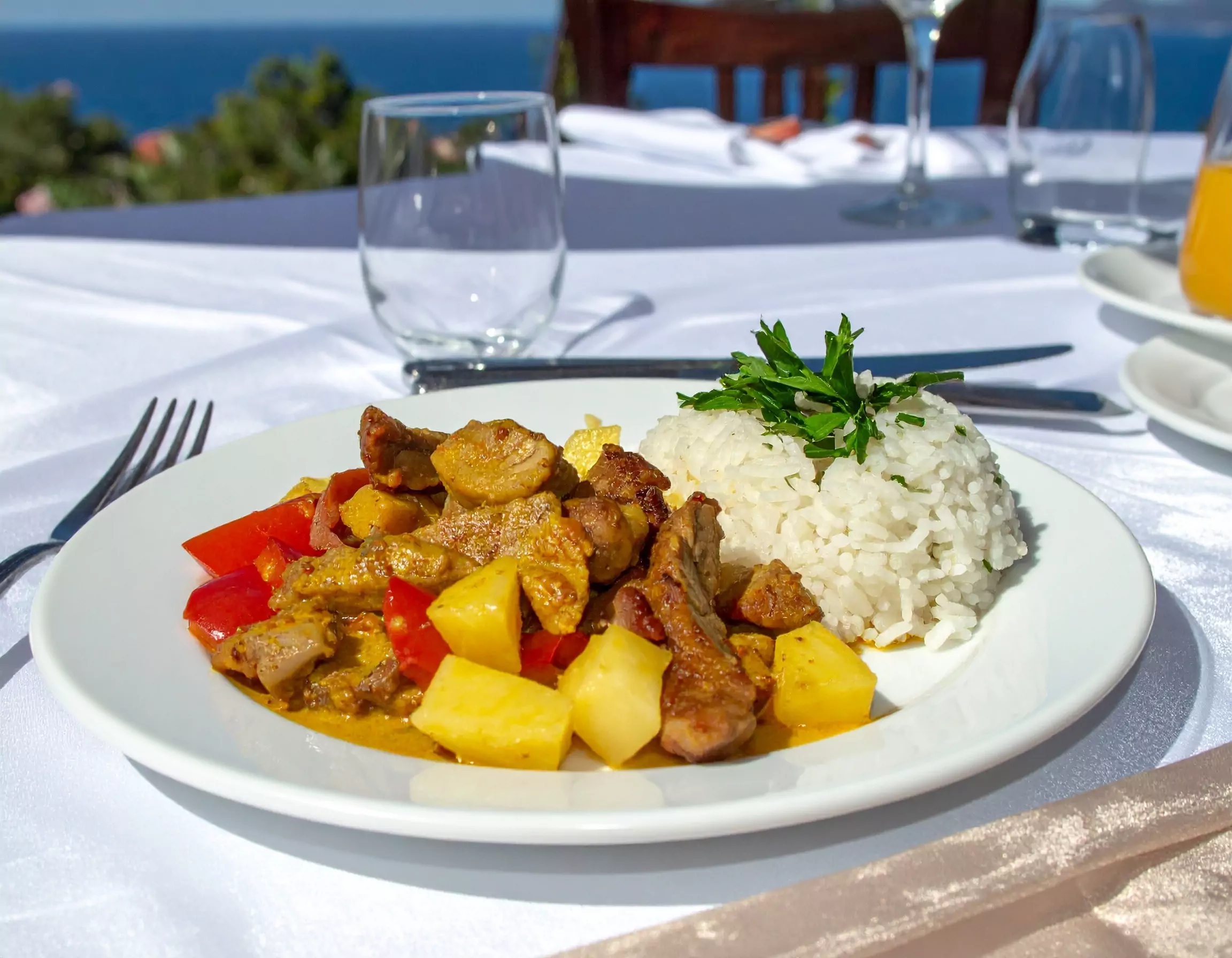 Teller mit Reis garniert mit Petersilie, gelber Curry- oder Saucengericht mit gebratenem Fleisch, Ananas und roten Paprikastücken, auf einem weißen Tisch mit Messer, Gabel und leerem Glas, im Freien bei Sonnenschein mit Meerblick im Hintergrund