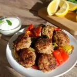 Weißer Teller mit gebratenen Frikadellen auf gegrilltem buntem Gemüse, daneben eine Schale mit weißer Soße und Minzblättern, im Hintergrund ein Schneidebrett mit Zitronenspalten auf einem Holztisch.