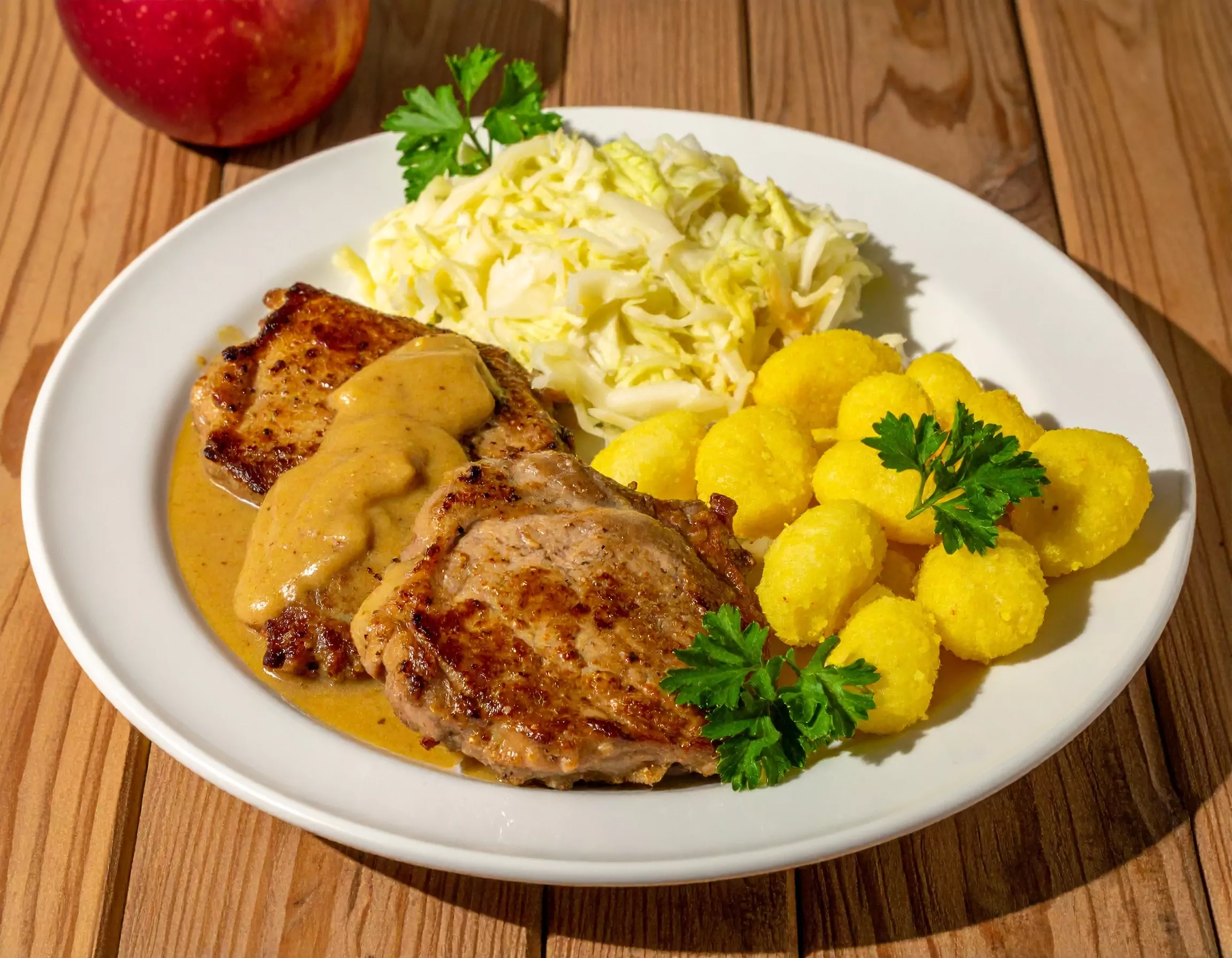 Weißer Teller mit gebratenem Fleisch in hellbrauner Sauce, gelben Kartoffelknödeln, Krautsalat und Petersilienzweigen, auf Holztisch mit rotem Apfel im Hintergrund