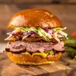 Burger mit geräuchertem Rindfleisch, Senf, Gewürzgurken, Rotkohl, Zwiebeln und Krautsalat in einem glänzenden Brioche-Brötchen auf einem Holzbrett vor unscharfem Hintergrund