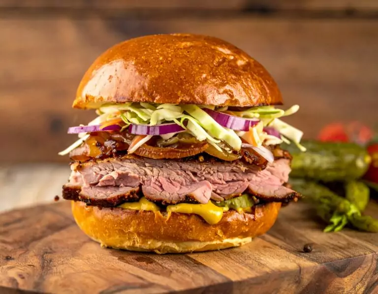 Burger mit geräuchertem Rindfleisch, Senf, Gewürzgurken, Rotkohl, Zwiebeln und Krautsalat in einem glänzenden Brioche-Brötchen auf einem Holzbrett vor unscharfem Hintergrund