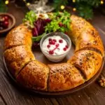 Runder, goldbraun gebackener Brotring mit Sesam bestreut, in Segmente geschnitten auf einem Holzbrett, in der Mitte eine Schale mit weißem Dip und roten Beeren, daneben frische grüne Salatblätter und rote Beeren, im Hintergrund Holzoberfläche und grüne Zweige